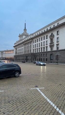 The video shows a wide shot of a city street paved with yellowish-brown cobblestones, appearing wet and reflective, suggesting recent rain or damp conditions. White painted markings are visible on the cobblestones in the foreground.

In the background, a very large, imposing building dominates the scene. It features a classical architectural style with multiple stories. The upper facade is light-colored (white or very light grey), while the lower levels are constructed from a darker, grey stone. The building has numerous rectangular windows, some of which are adorned with small balconies. A distinctive tower or spire with a pointed top rises from the roof, slightly to the left of the building's center. Further down the street, another large, similar-looking building can be seen in the distance on the left.

Several cars are present in the video. In the immediate foreground, on the left, the rear and side of a black SUV are visible, seemingly parked or moving slowly. In the middle ground, a silver sedan drives from the left side of the frame towards the right, its headlights subtly reflecting on the wet ground. Other vehicles, including a few with headlights on, are visible further down the street, moving in various directions.

The sky is overcast and appears to be either early morning or late afternoon/evening, as the light is diffused and soft. The overall ambiance is somewhat subdued due to the weather conditions. The architecture of the main building strongly suggests a location in Eastern Europe, reminiscent of the Largo complex in Sofia, Bulgaria, specifically the former Party House (now National Assembly).