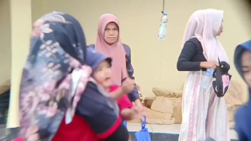 A group of people are gathered outdoors, possibly near a building. In the foreground, a person wearing a black hijab with a floral pattern is partially visible. Next to them, a child wearing a blue hat and a red shirt is being carried. The child is holding a stick. To the left of the child, another child with short hair is looking towards the left.

Behind these individuals, a woman in a pink hijab is looking towards the left. Further back and to the right, another person in a light-colored hijab is holding a bottle of water and a bag. In the background, there are stacks of what appear to be sacks or bags. A bottle of water is hanging from a wire or string. The overall lighting suggests it is daytime.