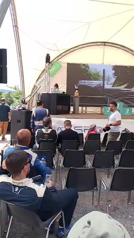 A group of people are gathered in front of a stage under a large white tent. The stage has a screen displaying a racing scene, a podium with numbered places, and several speakers. Several people are seated in chairs facing the stage, and a few individuals are standing behind the stage. A red and white checkered archway with "MA-FRA" printed on it is visible in the background, as is a building with multiple balconies. The ground is covered in gravel, and there are trees and foliage visible around the area. Some people are wearing racing suits.