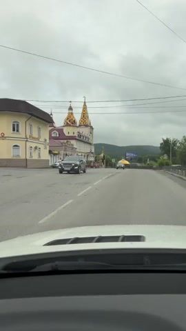 A video captures a view from inside a vehicle driving on a cloudy day past a building with distinctive yellow, patterned spires and red roofs. The building has a light-colored facade with arched windows and decorative elements. On the left side of the road, there is a smaller yellow building. Several cars are visible on the road. One gray sedan is parked on the left, and a black SUV is driving on the road. Further down the road, other vehicles, including a white van and a light-colored car, are visible. The road appears to be paved, and there are power lines overhead. In the background, hills can be seen under a gray, overcast sky.