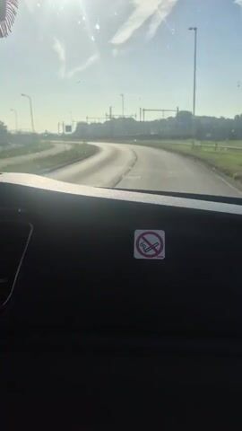 The camera faces forward from inside a vehicle, looking through the windshield. The dashboard is visible in the lower portion of the frame. A "no smoking" sign is affixed to the dashboard. Outside the windshield, a paved road winds through a landscape with sparse greenery and a light blue sky with some clouds.  A few light poles line the roadside.  A bridge-like structure is seen in the distance. A car travels on the road in the distance. The sun shines brightly, partially obscured by clouds, creating glare on the windshield. A decorative hanging item, possibly with a flag design, is briefly visible near the top of the windshield.