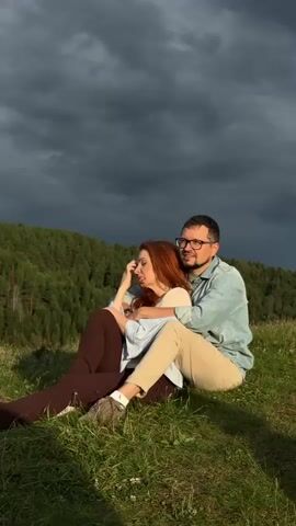 A man and a woman are sitting on a grassy hillside. The man has his arms around the woman, and she is leaning her head on his shoulder. They are both looking to the left. The background is a dark, cloudy sky and a forested hillside. The video is shot from a low angle, with the couple in the foreground and the landscape behind them. The lighting is dim, with the sun casting some light on the couple.
