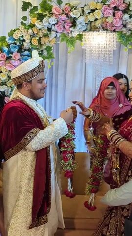 The video shows a wedding ceremony. The groom, wearing a white and maroon outfit and a traditional hat, is standing and smiling as he lifts the bride, who is wearing a maroon and gold sari and jewelry. People are around them, some are cheering and taking pictures with their phones. The background is decorated with flowers and a chandelier.
