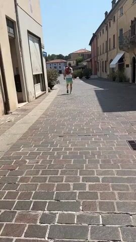A person is walking down a narrow street paved with cobblestones. The person is wearing a red shirt and white shorts. The street is lined with buildings on both sides, and there are trees and plants along the sides of the street. The sky is clear and blue. The sun is shining, casting shadows on the cobblestones.