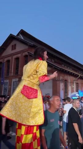 This is a vertical video showcasing an outdoor event, likely a festival or gathering, taking place in Kragujevac, Serbia, during dusk. The sky is a clear blue, transitioning to a soft orange hue near the horizon, indicating the setting sun.

The primary subjects are people enjoying the event and performers on stilts. In the foreground, a person in a vibrant yellow and red costume, adorned with elaborate patterns and a red pocket, is walking on stilts. Their costume is theatrical and eye-catching. Further into the crowd, another performer in a jester-like costume, featuring a red and black diamond pattern and a pointed hat, is also on stilts, interacting with the attendees.

The scene is bustling with a diverse crowd of people of various ages, dressed in casual summer attire. Many are looking towards the performers or socializing with each other. Some are holding drinks, suggesting a relaxed and festive atmosphere. In the background, there are glimpses of event infrastructure, including a stage or platform, tents with red canopies, and a large brick building that might be an industrial structure given the presence of a tall smokestack in the distance. The overall mood appears joyful and celebratory, with people engaged and seemingly having a good time.

There is some visible text on a red banner in the background, appearing to say "VIP 3" and possibly other event-related words which are not clearly legible. The presence of a moon in the sky further confirms it is evening.