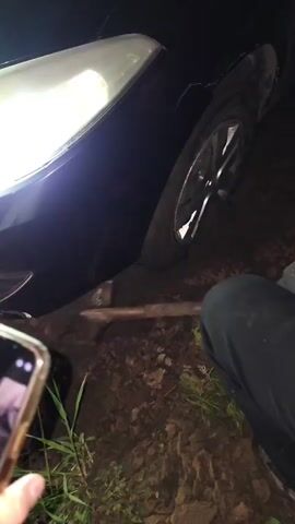 A close-up shot shows the front right side of a black car with its headlight on. The car's tire is partially submerged in dirt and mud, suggesting it may be stuck. A shovel is positioned in the dirt near the tire, with its handle extending towards the right side of the frame. The ground is uneven and appears to be a natural, unpaved surface. The lighting suggests it is nighttime or a very dark environment. The footage is shaky and appears to be handheld.