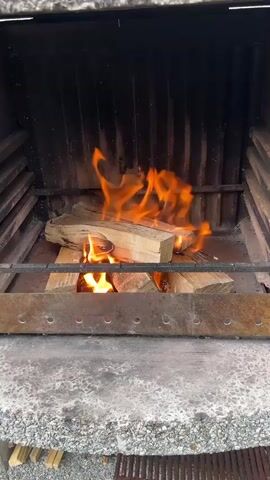 A video shows a close-up of a fire burning in a grill. The flames are bright orange and yellow, and they lick at the pieces of wood stacked on the grate. The grill itself appears to be made of stone or concrete, with metal bars for the grate. The background is dark, suggesting the grill is in an outdoor setting, possibly at night or in a shaded area. There are no people or other objects visible in the frame besides the fire and the grill. The video is short, lasting only a few seconds, and captures the dynamic movement of the flames.