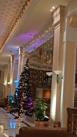 The video shows a decorated hotel lobby. A decorated Christmas tree sits to the left of a wall of decorative string lights hanging from the ceiling. The lobby is accented with columns, indoor plants, and lamps. A portion of a brown leather chair is visible at the bottom of the frame. A person walks by in the background.