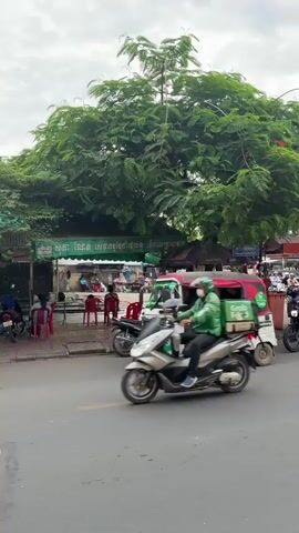 A street scene in a bustling urban environment, likely in Southeast Asia, unfolds under an overcast sky. A tuk-tuk, painted white and red with green accents and the "Grab" logo, moves through the frame from left to right. Several motorcycles and scooters navigate the road, some carrying passengers. Pedestrians are visible, some walking along the side of the road, others sitting near street stalls. Lush green trees provide shade over parts of the scene. A prominent sign is displayed on a pole, featuring a red circle with a slash through a drone icon, accompanied by the text "NO DRONE ZONE" in English, and Khmer script above. The background shows buildings, more stalls, and a general sense of activity, suggesting a commercial or market area. The lighting is diffuse, typical of a cloudy day.