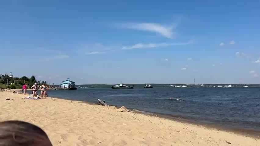 The video opens with a wide shot of a sandy beach on a sunny day. In the distance, a large body of water stretches to the horizon, dotted with several boats. Two ferries are prominently visible in the middle ground, moving across the water. On the left side of the frame, a person in a bikini is standing on the beach. In the water, a swimmer is visible, with their head and arms breaking the surface. The sky is a clear blue with a few wispy clouds. The shoreline is lined with trees in the distance, and a small building with a blue roof is situated on the left bank of the water. The scene appears to be a recreational area during daylight hours.