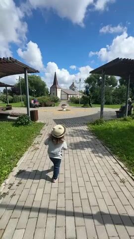 A young child wearing a straw hat and jeans walks down a paved pathway in a park. The child is seen from behind, heading away from the camera. The pathway is lined with green grass and trees. In the background, a building with a tall, pointed spire is visible, suggesting it could be a church or a similar structure. There are also pergolas on either side of the pathway, providing shade. The sky is a vibrant blue with fluffy white clouds, indicating fair weather and likely daytime. The scene appears peaceful and idyllic. There is no visible text in the media file.