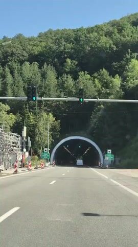 A car is driving down a paved road towards a tunnel entrance. The tunnel is large and arched, with lights illuminating the interior. Green traffic lights are visible above the road, indicating that it is safe to enter the tunnel. The road is lined with trees and foliage, and the sky is clear and blue. The car continues to drive into the tunnel.