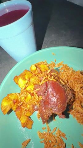 The scene is a close-up shot of a teal plate of food accompanied by a blue plastic cup filled with a dark red liquid. The food consists of reddish-orange rice, fried plantains, and a piece of dark, glazed meat. A hand holding a spoon scoops some of the rice off the plate. The food sits on a dark, possibly black, surface.
