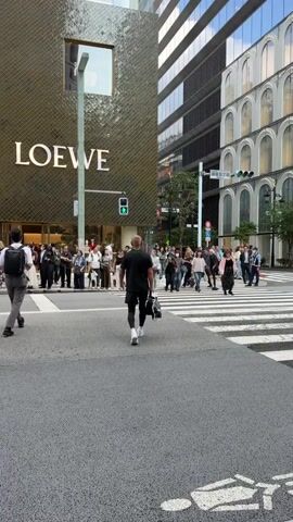The video shows a street scene with a crosswalk in the foreground. A man wearing a black t-shirt and black pants walks away from the camera towards the crosswalk. To the left of the man, a building with the "LOEWE" logo is visible. A group of people are standing and walking on the sidewalk and across the street. The traffic light is green. The scene appears to be in a city with modern buildings.