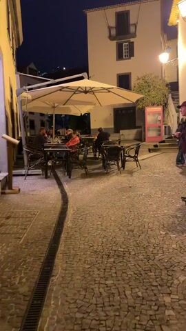 The video shows a narrow European street at night. The street is paved with cobblestones, and there is a drainage ditch running along the edge of the road. Several tables and chairs are arranged on the street outside a restaurant, and some people are sitting at the tables, presumably dining. A large white umbrella provides shade for some of the tables. Across the street, there is a tall, light-colored building with a balcony. A pink phone booth stands near the building. Two people are standing on the sidewalk near the edge of the frame, engaged in conversation. The street is illuminated by streetlights.