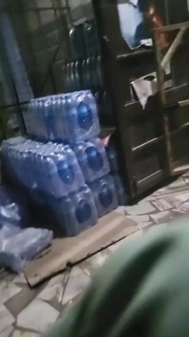 The video begins with an indoor shot of a storefront, the camera moving towards the entrance. The camera then moves to an outdoor scene, passing a stack of wrapped items, potentially cases of bottled water. It continues past parked vehicles, then pans to show more of the wrapped items stacked outside, a motorcycle parked behind a chain link fence, and additional parked vehicles. The overall impression is of a commercial area with goods being transported or stored.