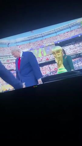 A man in a dark suit shakes hands with a man in a blue suit on a stage. There are two other people on the stage, one in a green dress and one in a dark suit. In the background, a stadium is filled with spectators. A golden trophy is visible on a pedestal to the right of the stage. The man in the blue suit turns and walks away, followed by the man in the dark suit.