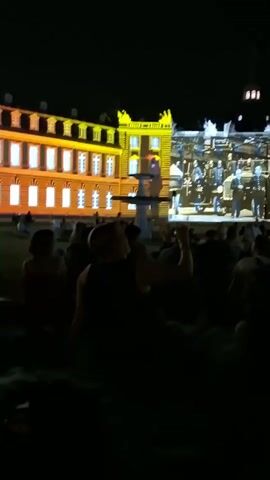 The video shows a nighttime outdoor event where a building facade is illuminated with a projected light show. The building appears to be a historic structure with multiple windows and architectural details, some of which are highlighted in yellow. The light show features images of people, possibly historical figures in uniform, arranged in rows and columns across the facade. In the foreground, a crowd of people is seated on the ground, facing the building and watching the display. The overall atmosphere is dark, with the projected light being the primary source of illumination for the building and the scene.