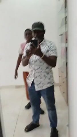 A man is holding up his cell phone and taking a selfie in front of a large mirror. In the mirror behind him, a child stands and poses with their arms raised in peace signs. To the right of the mirror, a glass cabinet displays a collection of purses. The man is wearing a light-colored button-down shirt with a pattern, blue jeans, and a dark-colored cap. He also has a beard and is wearing a bracelet. The child is wearing a t-shirt and brown pants.