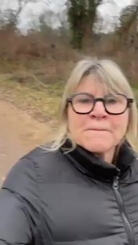 A woman is taking a video while walking on a dirt path through a wooded area. The woman has blonde hair, glasses, and is wearing a black puffer jacket. The trees in the background are mostly bare, and there is some green grass and brown foliage along the sides of the path. The sky is overcast. The woman smiles and speaks to the camera, saying she is photographing the landscape for her son.