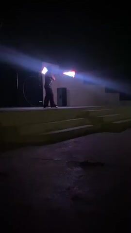 A person is juggling illuminated objects on a dimly lit stage at night. The stage has multiple levels, and a spotlight shines on the performer, who is dressed in dark clothing. The illuminated objects appear to be square or rectangular and display multiple colors. The background is dark, with some indistinct structures visible behind the stage. The music plays with a techno rhythm.