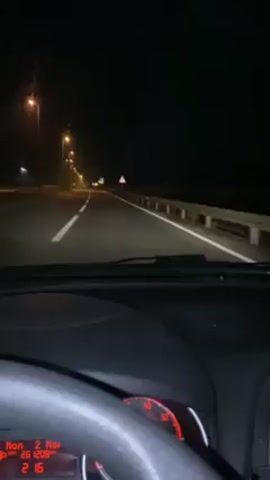 The video shows a nighttime scene from inside a car driving on a highway. The road is illuminated by streetlights, and the dashboard of the car is visible. There are road signs along the highway. The car appears to be moving at a steady speed.