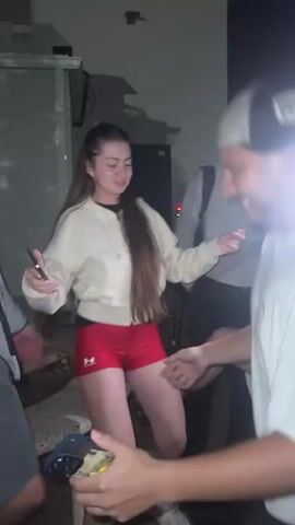 A group of five young adults, appearing to be in their late teens or early twenties, are gathered outdoors at night. The lighting is dim, with some artificial light sources casting shadows. A young woman in the center, wearing a beige hoodie and red athletic shorts, is dancing with her arms extended. She is smiling and looking towards a young man in a white t-shirt and a baseball cap who is facing her and also smiling. Another young man in a grey t-shirt and shorts is to her left, also smiling and gesturing with his hands. To the far left, a young man in a grey hooded jacket and shorts is visible, his hands held out. Another young man is seated in the foreground on the right, partially obscured by the others, wearing a white t-shirt and a baseball cap. The background shows a building with an orange awning and a dark sky. The overall atmosphere suggests a casual social gathering or party.