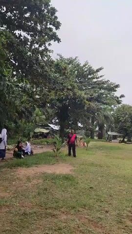 In an outdoor grassy area, a person in an orange vest and black pants is preparing to throw something. Another person in a red shirt and black pants stands further away, facing the first person. A few other individuals are visible in the background, including some seated on the left side of the frame. Trees and a few structures are present in the background. The sky is overcast.