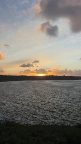 The video shows a sunset over a body of water. The sun is low on the horizon, casting a warm glow across the sky. There are a few clouds in the sky, some of which are illuminated by the sun. The water is dark and choppy, with waves visible. In the foreground, there is a patch of dark green vegetation, possibly bushes or trees, covering a slope. The overall scene is serene and picturesque, capturing the beauty of nature at dusk.
