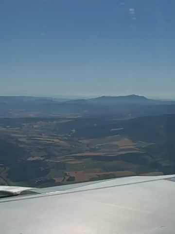 The video shows a view from an airplane window as the plane flies over a landscape. The landscape is a mix of hills, fields, and some distant mountains. The sky is blue and clear, with the sun shining brightly. Part of the airplane wing is visible in the foreground of each frame, extending from the bottom left corner of the image and tapering towards the right. In the first frame, the wing is a large, reflective surface. In subsequent frames, the wing continues to dominate the foreground. The landscape below appears to be a patchwork of agricultural fields in various shades of brown and green, interspersed with darker forested areas. The distant mountains are hazy blue shapes against the horizon. The sun's rays create lens flare effects as they stream through the airplane window, visible as bright streaks and circular patterns. The wing itself has visible panel lines and some small rivets or fasteners. The edge of the window frame is also visible in the later frames.