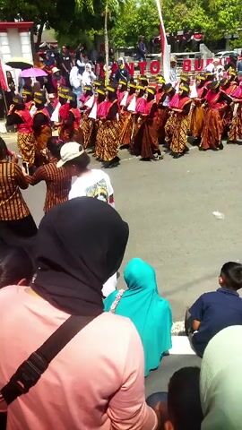 The video shows an outdoor event with a procession of people in traditional attire. In the foreground, the backs of several spectators are visible, including a person wearing a pink shirt and a black headscarf, and another wearing a teal headscarf. A child in a dark blue shirt is also seated. In the middle ground, a group of performers, mostly women, are lined up in rows, wearing colorful patterned skirts and tops, some with head coverings. They appear to be participating in a parade or performance. Behind them, more people are visible, some holding umbrellas and banners. The environment is an outdoor street, with trees and buildings in the background. The ground is paved. The overall scene suggests a cultural festival or celebration.