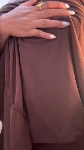 A young woman is posing in a brown dress with flowing sleeves. She has a gold ring on her left hand. Her hair is pulled up in a bun. She is wearing a backless dress with chains draping down her back. She turns to show the back of the dress. The background is a plain white wall.