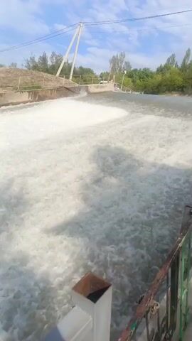 A video shows a body of water with white foam, likely from a dam or spillway, cascading down. In the background, trees line the horizon under a partly cloudy sky. A power line with poles runs across the scene. A metal railing runs along the bottom right of the frame, with a partial view of a white structure below it. The camera appears to be stationary, looking down at the turbulent water.