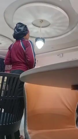 The video shows a woman in a maroon tracksuit and a black hair bonnet standing in a room. There are several people seated at a table, and the room has a modern ceiling design with a chandelier. The woman is facing the group at the table. The scene appears to be in a restaurant or a similar gathering place.
