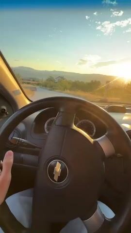 The video shows the interior of a car driving down a road during sunset. The camera is positioned behind the steering wheel, facing forward. The dashboard is visible, including the steering wheel, instrument cluster, and radio. The road ahead leads through a rural landscape with rolling hills and scattered houses. The sun is setting in the distance, casting a warm glow over the scene and creating lens flares. A hand is seen briefly touching the steering wheel. The sky is a deep blue with some clouds.