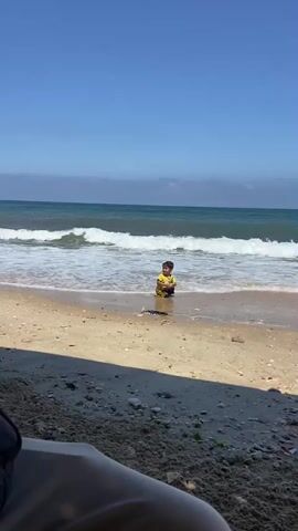 A young child wearing a yellow shirt is playing in shallow water at a beach. The child is facing the ocean, with waves breaking in front of them. The beach is sandy, and there are some pieces of debris on the sand in the foreground. The sky is clear and blue, and there are some clouds on the horizon. The video is shot from a low angle, possibly from someone sitting on the sand.