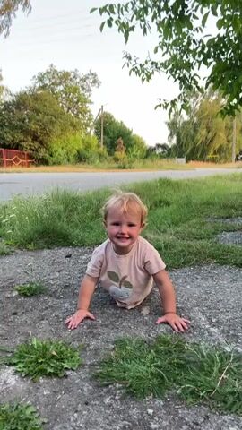 A blonde toddler wearing a light pink t-shirt with an apple design and beige pants is on the ground, crawling on the pavement with grass and trees in the background. The toddler smiles and looks at the camera, then turns their head away. The toddler then crawls out of frame.