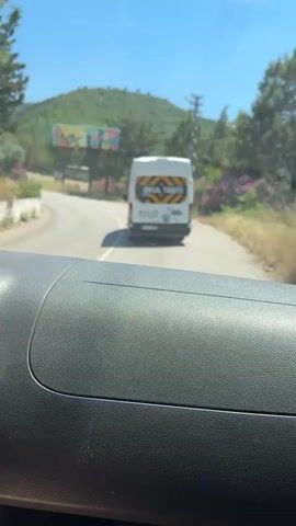 The video shows a view from inside a vehicle, looking forward and then to the side. In the forward view, a white van with yellow and black hazard stripes on the back is seen driving on a road. There is a billboard on the left side of the road in the distance. The road is curved and surrounded by greenery, trees, and hills under a bright blue sky. The view then shifts to the right, showing the interior of the vehicle's door and window. Outside the window, there are trees, flowering bushes with pink flowers, and a power line pole. The vehicle appears to be in motion on a rural road.