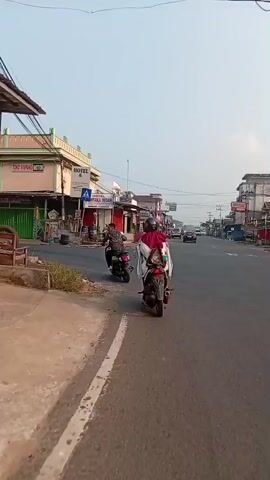 A first-person perspective video shows a rider on a scooter traveling down a street. The rider is wearing a dark helmet and a white jacket over a red garment, with a brown backpack. Another scooter is ahead and to the left. The street is lined with buildings, some of which appear to be shops. Several other vehicles, including a truck, are visible on the road. The sky is clear.