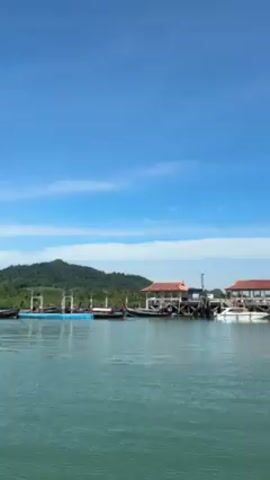 The video presents a serene view of a harbor on a clear, sunny day. The camera, likely positioned on a boat, floats on water, showing a line of boats of various sizes docked along two wooden structures that are both elevated over the water. The structures appear to be small buildings, possibly shelters or waiting areas. Behind these, a lush, green landscape stretches along the horizon, possibly representing an island or mainland. The sky is a brilliant blue, dotted with a few wispy clouds. The sea is calm, reflecting the sky and surrounding landscape. No people are visibly present, and no specific actions are occurring beyond the gentle movement of the water. The overall scene suggests a peaceful maritime setting.