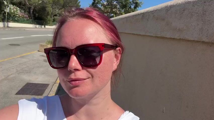 A woman wearing sunglasses and a white top stands outside next to a wall. She lifts the front of her floral skirt and places her hand inside.