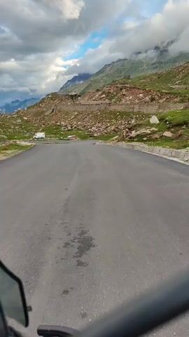 A first-person perspective video shows a motorcycle or car driving on a paved road through a mountainous landscape. The road curves to the right. On the left side of the road, there are grassy slopes dotted with rocks and some bushes. Further up the slope, there are more rocks and a winding road is visible, along with a white van. In the background, large mountains rise, with some parts obscured by clouds. The sky is partly cloudy.