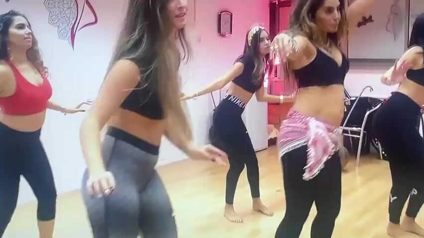 A group of five young women are in a dance studio. They are all dressed in athletic or dance attire. The woman in the foreground on the left is wearing a red sports bra and black leggings. To her right, a woman in a black crop top and grey and black patterned leggings is in the center of the frame. She is bending at the waist with her arms raised. Behind her, a woman in a black crop top and black leggings with "NIKE" written on them is also dancing. To the right of center, a woman wearing a black sports bra, black leggings, and a pink sequined belt is dancing. The woman furthest to the right is wearing a black crop top and black leggings. All of the women appear to be performing a choreographed dance. The room has white walls with some artwork and text on them, and a wooden floor. There is also some studio equipment visible in the background, including what looks like a bicycle with a large hoop attached to it.