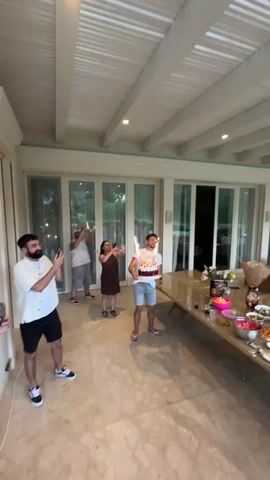 The video shows a group of people celebrating a birthday outdoors on a patio. A man in a white tank top and blue shorts is holding a birthday cake with sparklers that are lit. He is surrounded by other people, some of whom are taking pictures or videos of him with their phones. The patio has a tiled floor and a white ceiling with exposed beams. There is a long table laden with food and drinks in the background. The atmosphere appears festive and joyful.
