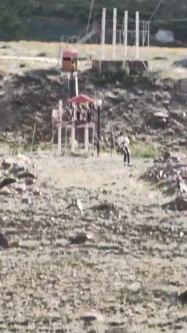 A zipline setup is visible on a rocky, grassy hillside. Several people are standing on platforms and one person is actively ziplining across the frame. In the background, a large mountain range and a turbulent river are visible.