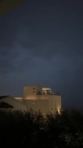 The media file shows a nighttime scene in Trap, Montenegro, featuring a modern, light-colored building with illuminated windows and rooftop seating. The sky is dark and cloudy, suggesting a stormy or overcast evening. In the foreground, dark silhouettes of trees and foliage obscure the lower part of the scene, creating a sense of depth and framing the building. The lighting on the building is warm and inviting, contrasting with the dark sky and the surrounding natural elements. There is no visible text in the media file. The overall mood is atmospheric and somewhat dramatic due to the interplay of light and shadow, and the ominous sky.
