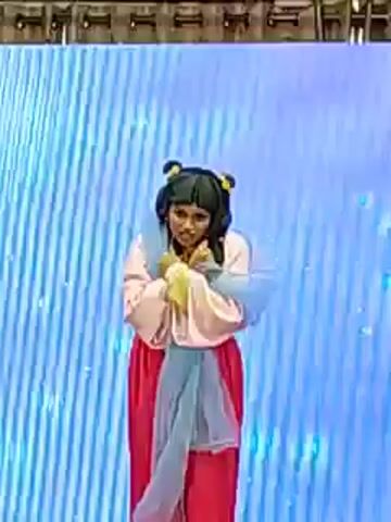 A person in an East Asian-style costume dances in front of a large blue LED screen. The dancer, who appears to be female, wears a light pink top, red skirt, and a pale blue sash. The hair is styled with traditional ornaments. The backdrop displays a grid pattern with varying light intensity. The lighting rig can be seen near the top of the frame.