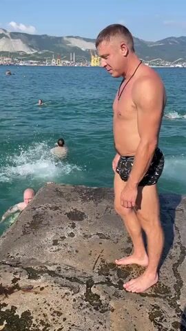 A man in black patterned swim trunks walks along a concrete pier towards the edge. He pauses, looks down at the clear turquoise water, and then dives in headfirst. The camera follows him as he splashes into the water and surfaces, looking up. In the background, other people are swimming, and a coastal town with mountains and industrial structures is visible. The sky is clear and blue.