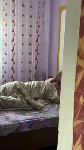 A person is shown sleeping in a bed, covered by a patterned blanket that appears to be a beige and grey animal print or camouflage design. The person is lying on their side with their face partially visible, eyes closed, suggesting deep sleep. Their head rests on a striped pillow featuring pink, purple, and white colors. The bed has a floral sheet with purple and green patterns.

The bedroom features light purple walls and white curtains adorned with red leaf or floral patterns. A wooden headboard is visible, and behind it, a golden-framed object (possibly a mirror or picture) can be seen. On the right side of the frame, a section of a wall or doorway is covered with floral wallpaper in shades of red, orange, and gold. The overall lighting suggests it is daytime, with natural light filtering through the curtains.