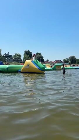 The video shows a busy lake on a clear day. In the foreground, the rippling, murky green water occupies most of the frame. In the middle ground, several inflatable water park structures are visible. One prominent yellow and blue inflatable slide has a person on top. Further back, other inflatable obstacles and a crowded beach with many people sunbathing and swimming are visible. The background consists of a clear blue sky and a distant tree line. The overall impression is of a lively summer day at a popular water recreation area.